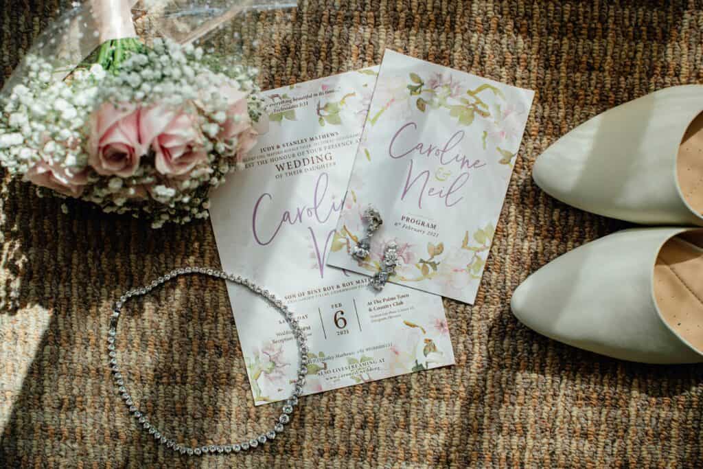 A wedding invitation from a high quality print shop surrounded by a bouquet of flowers, jewelries and lady's shoes. Zippy Printz