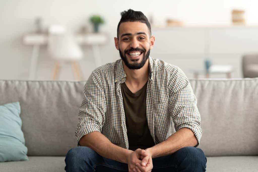 Portrait of handsome young Arab man smiling and looking at camera, sitting on sofa at home
